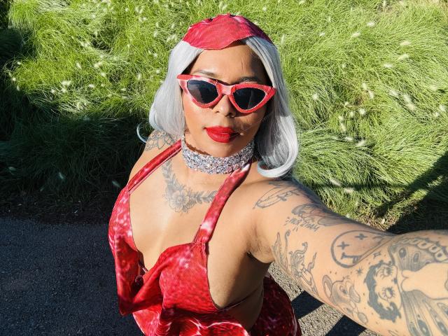 Chili with white hair, red dress, red sunglasses, red lips, white choker, tattoos, selfie, sunny day, green bush background.
