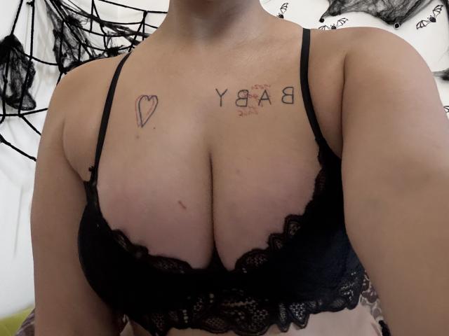 Les seins de Kametia, visibles sous un soutien-gorge noir en dentelle, présentent un tatouage en forme de cœur sur le côté gauche et "Y8AB" sur le côté droit. Sa peau est claire, et sa tête est hors du champ de vision..