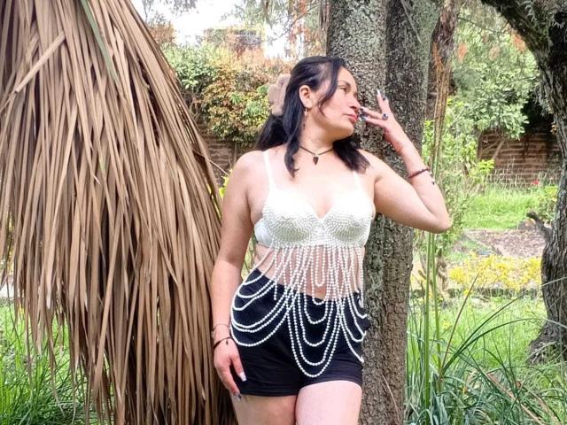Lily, de aspecto maduro, con cabello oscuro, una blusa de crochet blanca con cadenas de perlas, pantalones cortos negros, de pie junto a un árbol, fumando, mordiéndose el labio, mirando hacia un lado, al aire libre..