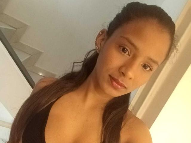 AriVelvet has brown skin, long dark hair in a ponytail, and is wearing a black top, showing her small tits and a subtle smile. She's looking at the camera.