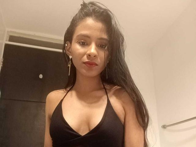 AriVelvet has long black hair, wearing a black halter top, showing her small tits, with a nose piercing, red lips, and a serious expression in a bathroom.
