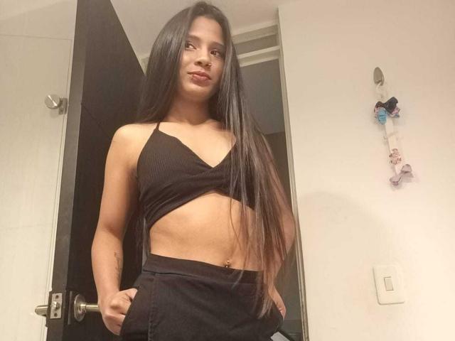 AriVelvet with long black hair, wearing a black crop top and matching black pants, hands in pockets, standing in a doorway, showing a flat stomach and belly button.