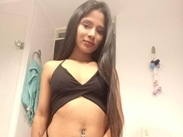 AriVelvet with long black hair, nose ring, wearing a black crop top, exposing her small tits and belly button piercing, smiling in a bathroom.
