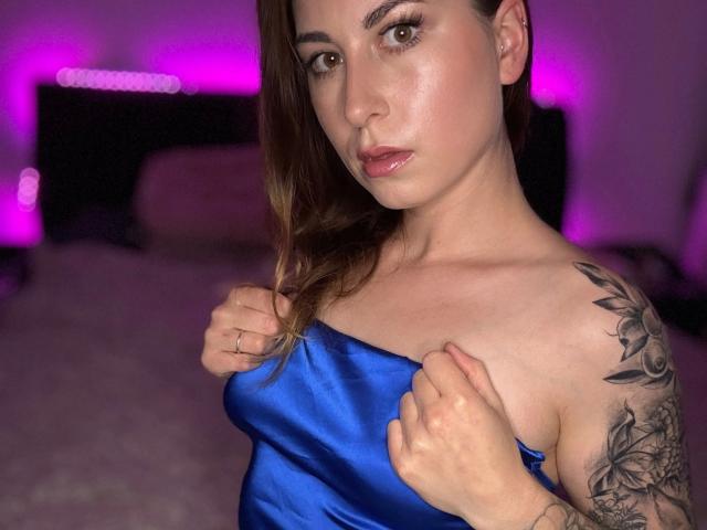 Mrsqueeniii-hot with shiny blue satin top, holding it against her tits, showing a tattoo on her right shoulder, and a serious expression, with pink lighting in the background.