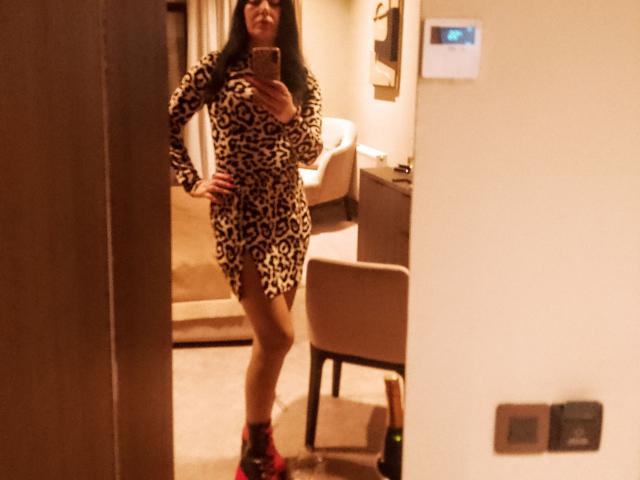 A blurry selfie of MirandaHott69 standing in a hotel room. She wears a leopard print dress with long sleeves, exposing her thighs, and red and black sneakers. Her right hand is on her hip, and she holds a phone in her left hand, partially covering her face. The room has a beige chair, a small table, and a light-colored carpet. The mirror reflection is slightly angled, capturing her from the side. The lighting is warm.