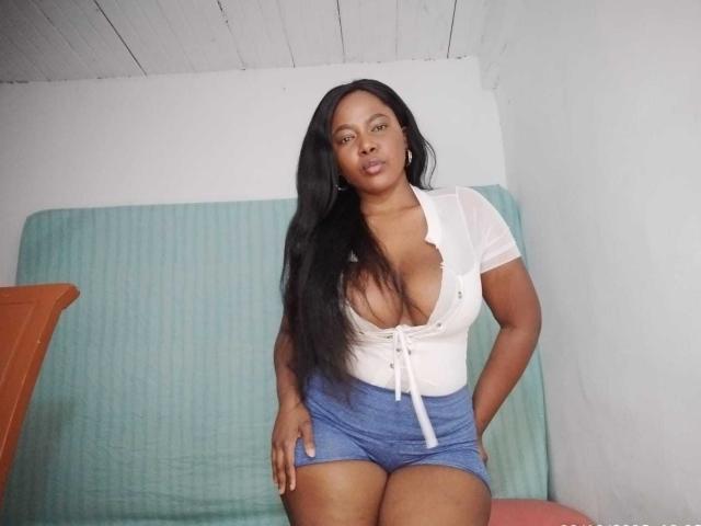MacarenaBoobs with long black hair, white deep-cut shirt showing big tits, and blue shorts, standing, hands on hips, looking at camera, in simple room with green fabric.