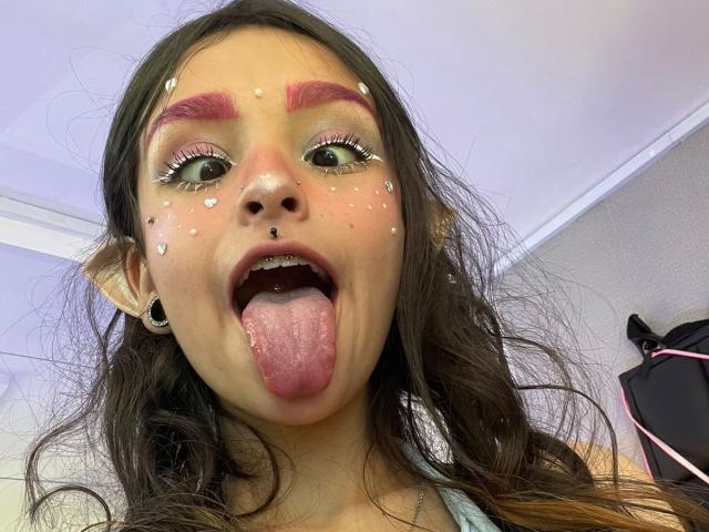 MarietteJones has pink eyebrows, white face paint, a nose ring, and black ear holes. She's sticking out her tongue, with long brown hair and green eyes.