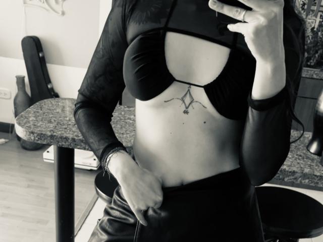 SujannaVarma's tits are in a black bra with a cutout, showing a geometric tattoo below. She's pulling down her pants, revealing a flat stomach and a black long-sleeve top.