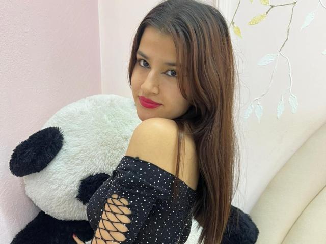 Iris Blair, met lang bruin haar, rode lippenstift en een zwart top met kanten details en een open schouder, omarmt een enorme witte panda, waarbij haar achterwerk zichtbaar is, in een zachtroze kamer..