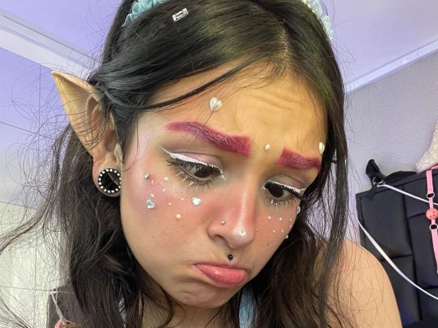 MarietteJones with pink eyebrows, elf ears, white face gems, black nose ring, and pink lipstick, looking down with pouty lips, wearing a blue headband, and black ear plugs.
