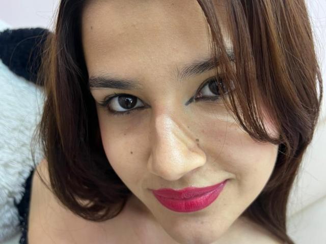 Iris Blair heeft bruine ogen, rode lippenstift en bruin haar van gemiddelde lengte met een lichte scheiding in het midden, en ze glimlacht naar de camera met een klein beauty markje op haar neus..
