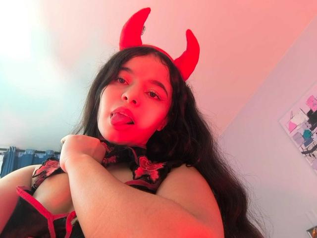 LissyCarter with red devil horns, long black hair, tongue out, wearing a black floral top, covering her tits with one hand, red and pink lighting, looking at the camera.