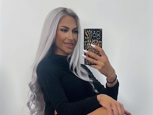 KarlaBart, con i suoi lunghi capelli argentati, indossa una maglia nera a maniche lunghe, sorride allo specchio, tiene in mano un telefono con una custodia dorata decorata, mostrando il suo seno e i suoi glutei nel riflesso..