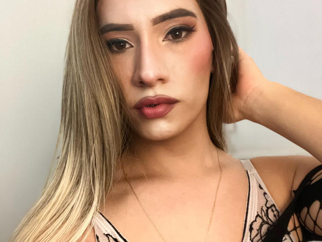 Close-up photo of CamiiNiieves with long, straight blonde hair, fair skin, and brown eyes. She has dark, well-groomed eyebrows, subtle pink blush, and pink lipstick. She's wearing a light pink lace top with black trim and a thin gold chain necklace. Her right hand is behind her head, slightly covering her ear. The image is focused on her face, with soft, even lighting highlighting her facial features. Her expression is neutral.