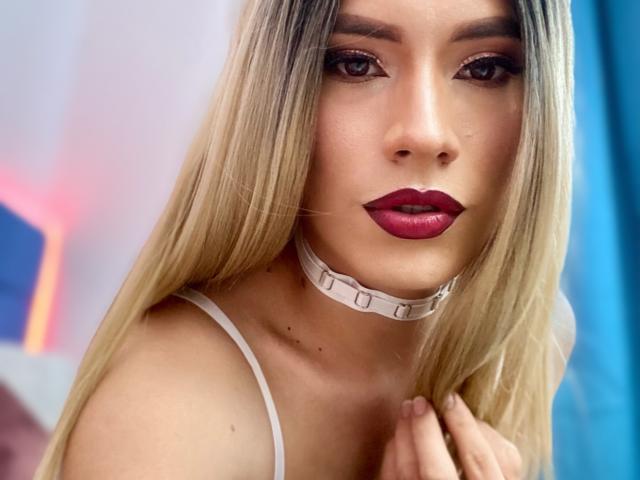 Close-up of CamiiNiieves with long, straight blonde hair and fair skin. She has dark, arched eyebrows, thick eyeliner, and dark red lipstick. She wears a white choker with "LOVE" in black letters and a thin white spaghetti strap. Her right hand touches her hair. The background has blue and pink neon light. The image focuses on her face and upper torso, highlighting her makeup and choker. Slightly blurred background emphasizes her presence.