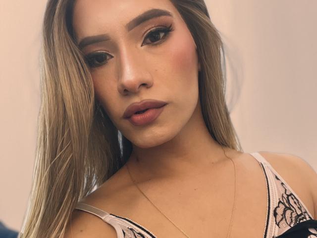 CamiiNiieves has long blonde hair, thick eyebrows, and pink lipstick. She's wearing a white lace top with black detailing, and a gold necklace. Her skin is light and freckled.