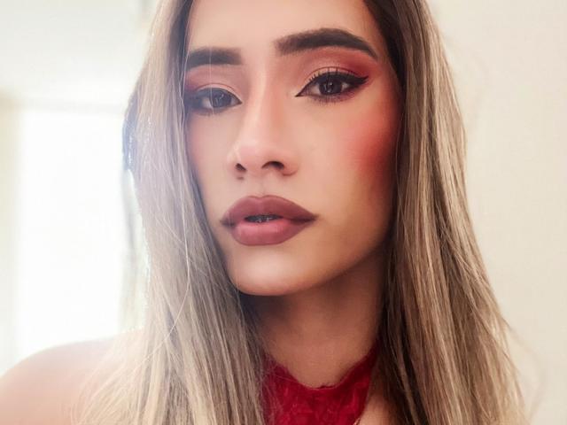 Close-up of CamiiNiieves with long, straight, blonde hair. She has a light skin tone, full eyebrows, and is wearing bold, winged eyeliner with pinkish-red eyeshadow. Her lips are full and painted a matte mauve. She has a slight, neutral expression. A red lace halter is visible around her neck. The image has soft lighting and a slightly blurred background. The composition centers on her face, highlighting her makeup and hair.