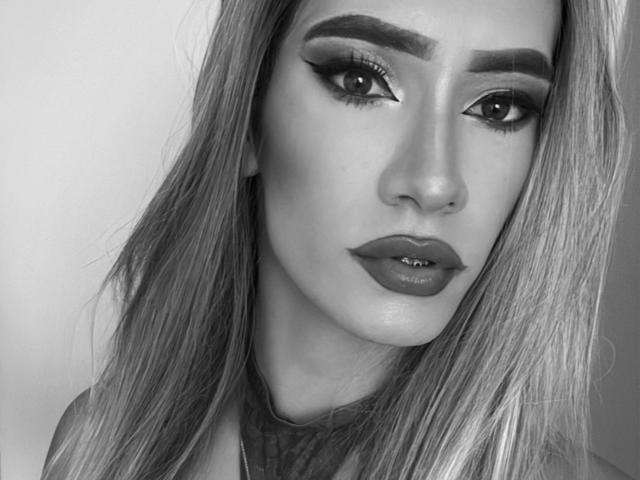 CamiiNiieves has glossy lips, thick eyebrows, and long hair. Her eyes are lined with dark eyeliner, and she's staring directly at the camera with a serious expression. Black-and-white photo.