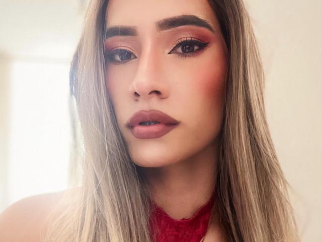 CamiiNiieves has long blonde hair, pink eyeshadow, winged eyeliner, red lipstick, and a red lace halter. Her face is slightly flushed with a serious expression.