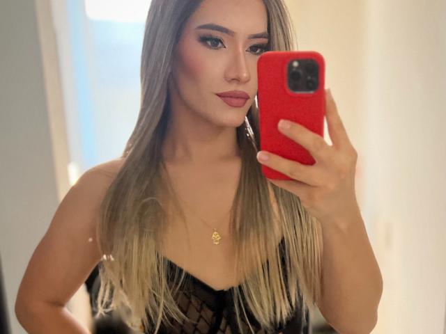 A selfie of CamiiNiieves with long, straight, blonde hair, wearing a black lace top. She holds a red phone in her right hand, taking the photo in a mirror. Her makeup includes bold eyebrows, dark eyeliner, and pink lipstick. She has a gold necklace with a small pendant. The focus is on her face and upper body, with a slightly blurred background. The image is sharp, with a warm, natural light. She has a confident expression.