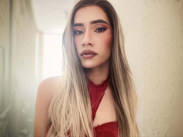 Close-up of CamiiNiieves with long, straight blonde hair and dark roots. She has prominent eyebrows, pink eyeshadow, and glossy pink lipstick. Her skin is light with a smooth texture. She's wearing a red lace halter top with a cutout at the chest. The background is softly blurred, with light and a textured wall. Her expression is neutral, slightly serious. The composition centers on her face, highlighting her makeup and hair. The lighting is soft, creating a gentle, almost ethereal effect.