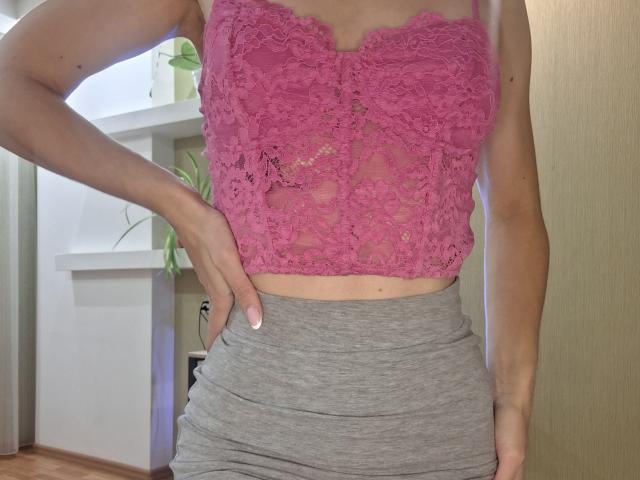 MilaYanis wearing a pink lace top with her tits barely covered, gray high-waisted skirt, hand on hip, showing her midriff and tan skin.
