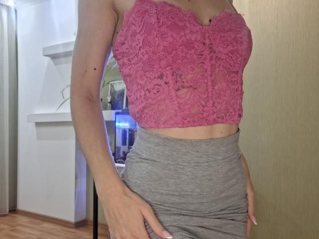 MilaYanis with pink lace bustier, showing her tits, and gray skirt, her hands on her hip, in a room with wooden floor and white shelves.
