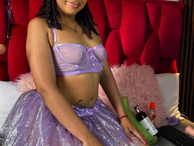 ArtemisaS with purple sheer bra, showing tits, and sparkly purple skirt, holding green dildo, bottle, and condom on bed with pink fluffy pillow. Dark curly hair, smiling.
