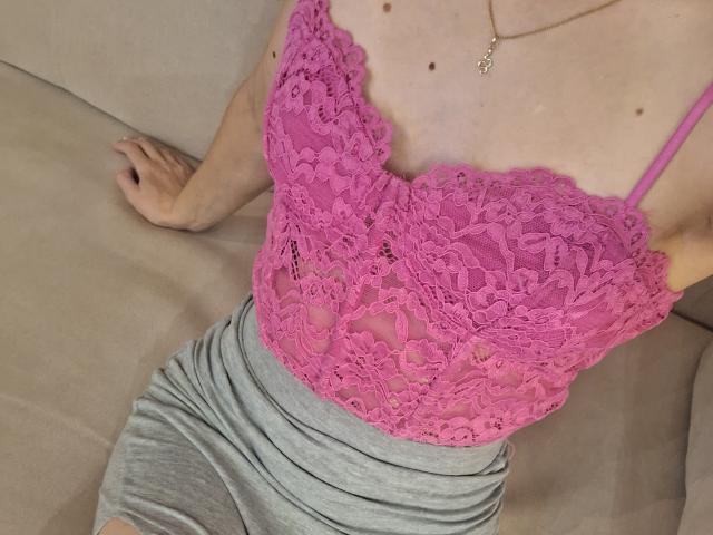 MilaYanis wearing a pink lace top that reveals her tits, gray skirt, and a gold necklace, sitting on a beige sofa.