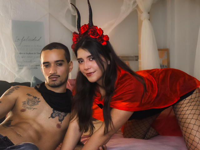 Laila Asher, met donker haar, een rode "devil" jurk en hoorns, leunt tegen een gespierde man zonder shirt, met tatoeages en een zwarte sjaal, en met visnetkousen, beiden zitten op een bed en kijken naar de camera..