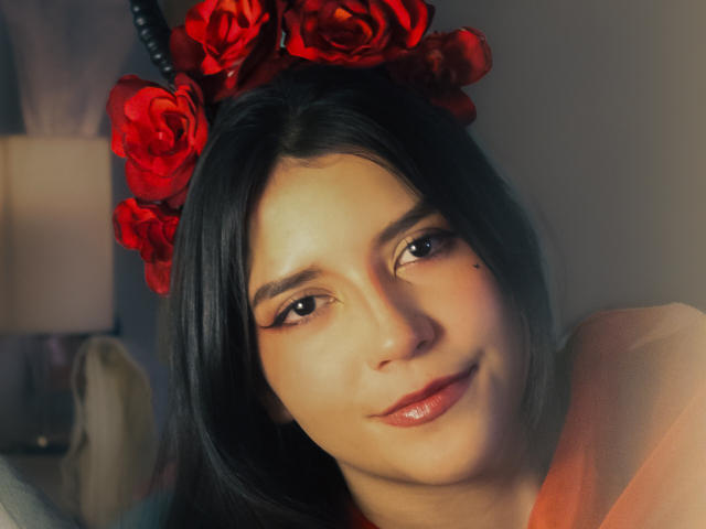 Laila Asher, met rode rozen in haar zwarte haar, glimlachend met glanzende, roze lippen, een kleine neus en eyeliner. Ze heeft een schattig, zacht uitziend gezicht, lichte huid en een subtiele glimlach..