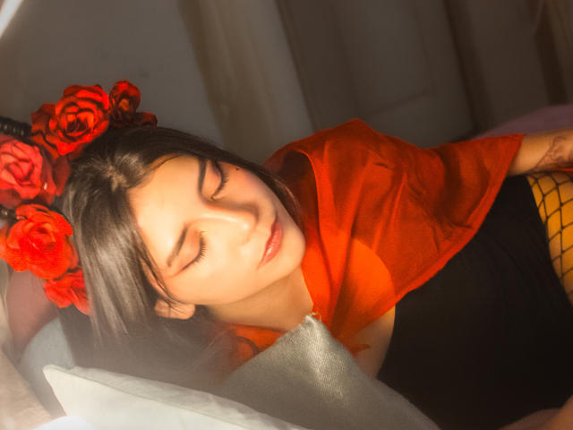 Laila Asher, con gli occhi chiusi, indossa rose rosse tra i capelli, una camicia arancione e un top a rete giallo, sdraiata su un letto, con il viso illuminato da una luce solare soffusa..