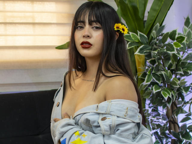 SopphieBloom with dark hair, sunflower in her hair, red lipstick, off-shoulder denim jacket, exposing her tits, standing by a plant, looking at the camera.