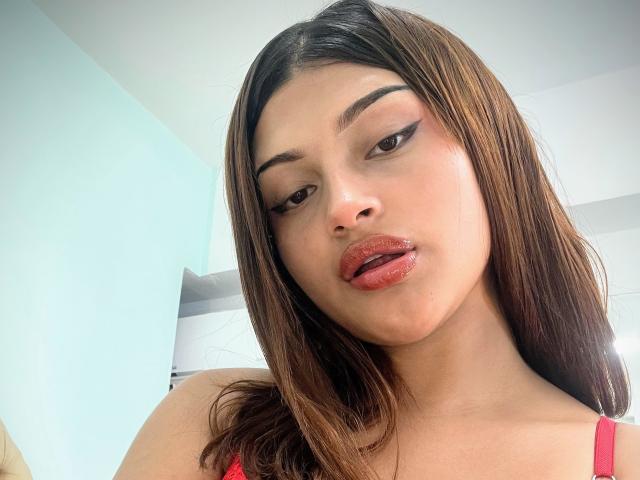 BrendaGreen has shiny brown lips, dark eyebrows, and straight brown hair. She's wearing red lipstick and a red top, looking at the camera with slightly parted, glossy lips.
