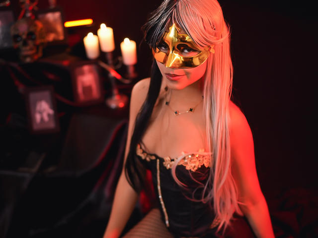 ChloeVixen with long white and black hair, wearing a golden mask, black corset with lace, and gold necklace, red lighting, candles in background, and a dark, mysterious vibe.