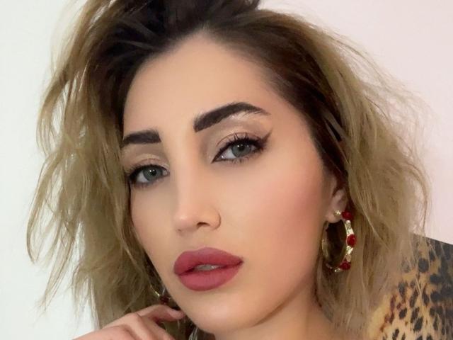 AngelsBack has a sexy face with full red lips, green eyes, and blonde messy hair. She's wearing hoop earrings with red beads and a leopard print top.