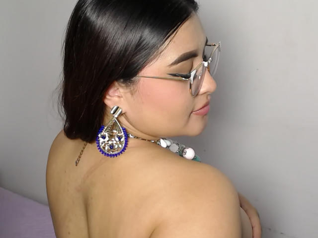 HelueJamil mit dunklem Haar, Brille und blauen, mit Schmuck besetzten Ohrringen, oben ohne, wobei ihr Rücken und Schulter zu sehen sind, lächelnd, wobei sie ihre Brüste mit ihrem Arm hält..