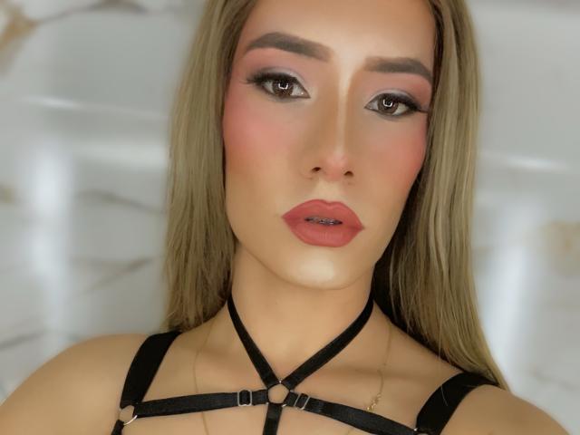 CamiiNiieves has pinkish-red lipstick, brown eyes with smoky eyeshadow, and blonde hair. She's wearing a black strappy harness with gold rings, looking directly at the camera.