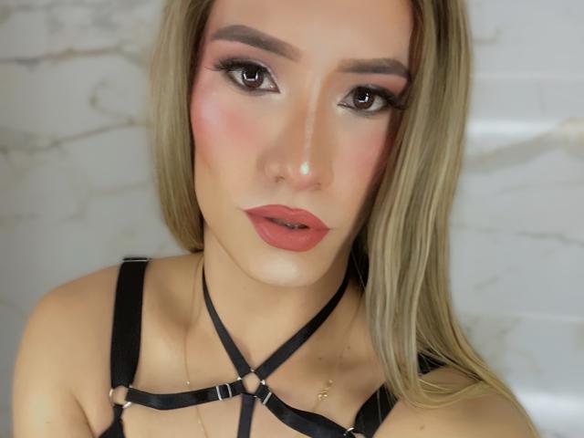 CamiiNiieves with blonde hair, pink blush, and red lipstick, wearing black harness, has brown eyes, and a neutral expression, looking at camera.