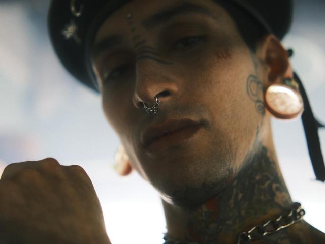Close-up of TonyoHatsumoto's face. He has a nose ring with a small decorative charm, large circular ear extensions, and multiple tattoos on his neck and chin. His skin is light, and he wears a black hat with a ribbon. His right hand is raised, slightly blurred. The background is out of focus, with cool blue and white hues. The photograph uses a shallow depth of field, emphasizing TonyoHatsumoto's facial features and tattoos. The image has a gritty, urban style.