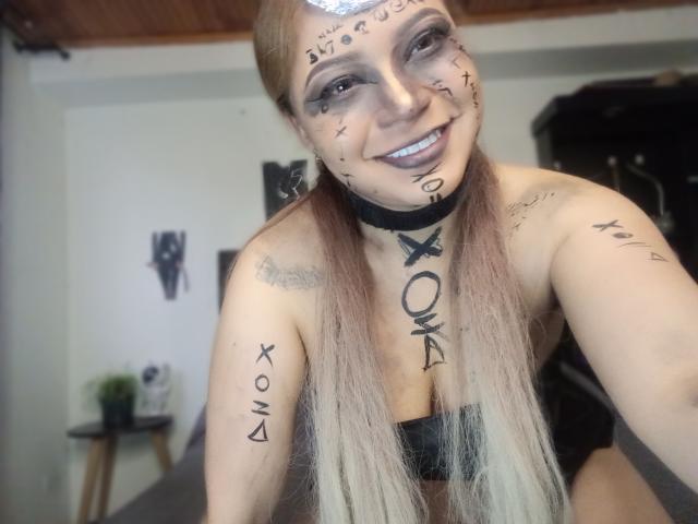 TatianaRossi with tattoos, long blonde pigtails, black choker, smiling, gray eyeshadow, black lipstick, small tits, arm tattoos, room with wooden ceiling, blurred background, smiling at camera.