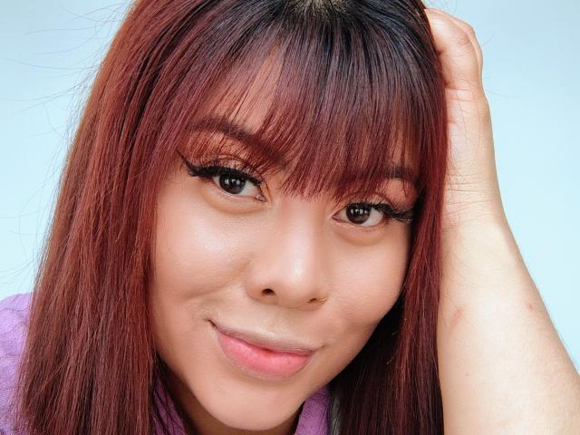 Noemi Thornee tiene el cabello rojo, pestañas gruesas y una ligera sonrisa, tocándose la cabeza con una mano, y lleva una blusa morada, con un piercing en la nariz en el lado derecho..