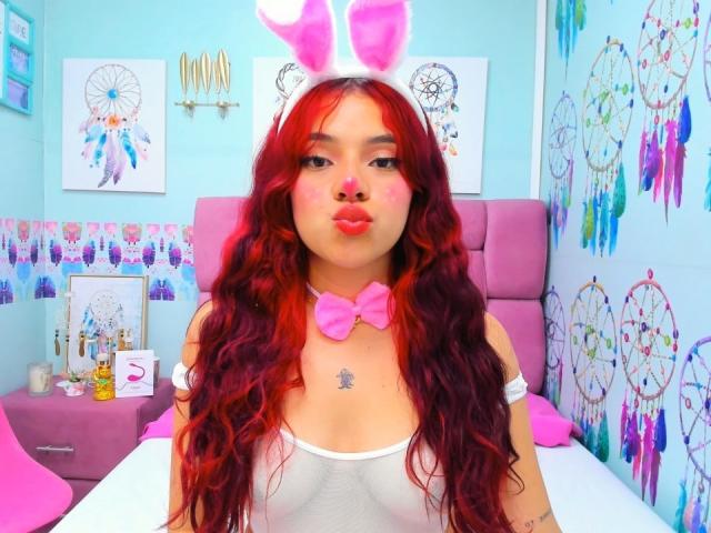 MiaLeroy, avec ses cheveux roux ondulés, ses oreilles de lapin roses, son débardeur blanc, son nœud papillon rose et son rouge à lèvres rose, faisant la moue, montrant ses seins et un petit tatouage au-dessus de sa poitrine..