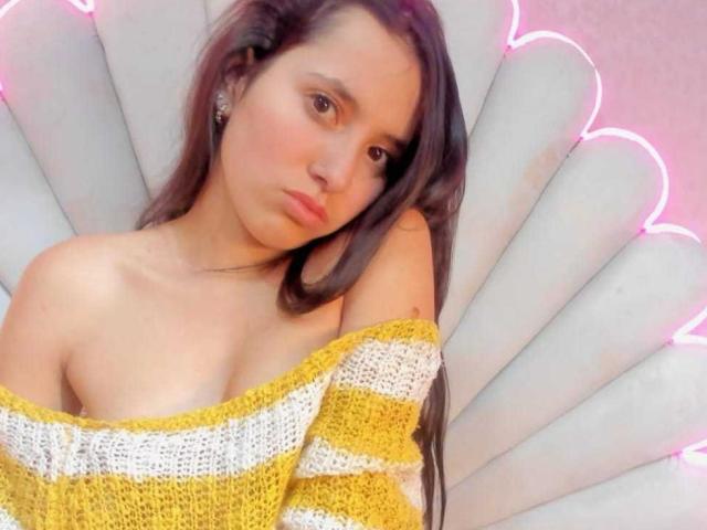 MallyBoom with long brown hair, yellow and white off-shoulder knit top, exposing small tits, sitting on white fan-shaped chair, pink neon lights in background.
