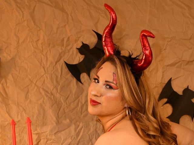 AlinaHill with red devil horns, black bat wings, and red lipstick, looking over her shoulder, showing her bare back and blonde curls, with red candles in the bottom left.