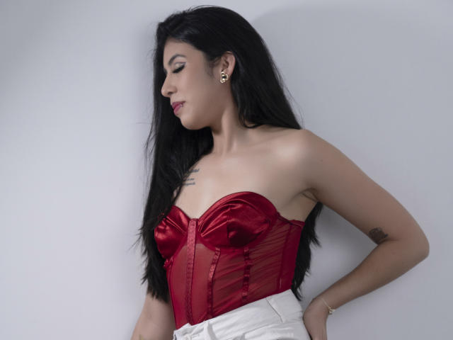MiluSky with long black hair, wearing a red satin bustier that accentuates her tits, and white pants, looking to the side with a subtle smile.