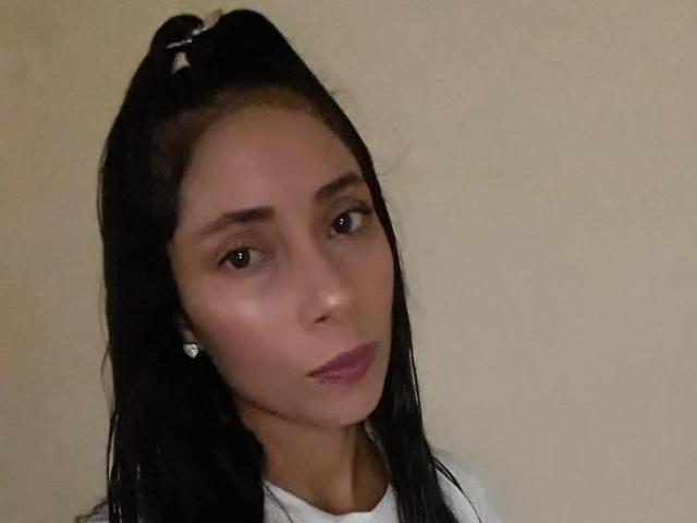 SaraySweetSex has dark brown hair in a high ponytail, brown eyes, light skin, and pink lipstick. She's wearing white and small diamond earrings.