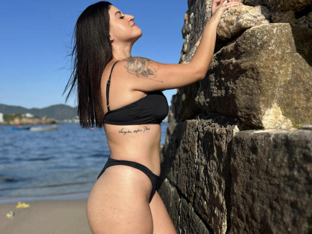 JuliaMenezes with black bikini, standing against rock, showing tattoo on right arm, hands on stone, eyes closed, long black hair, tan skin, visible ass, small breasts, tattooed side.