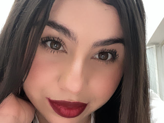 VenusxMusa with dark brown eyes, full red lips, and long brown hair, looking into the camera with slightly parted eyebrows and a subtle smile. Her eyelashes are long and dark.