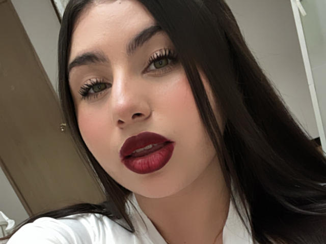 VenusxMusa has long black hair, green eyes, and full red lips. She's wearing a white shirt, looking at the camera with a slight pout. Her face is slightly flushed.